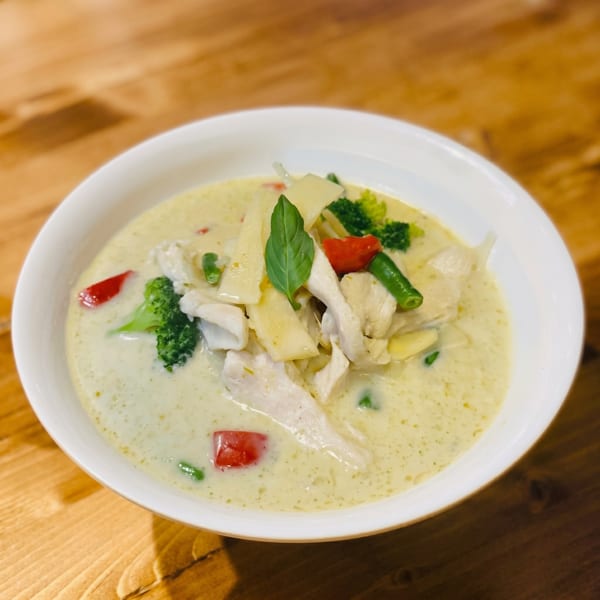 A white bowl of green curry with chicken, bamboo shoots, broccoli, red peppers, and basil.