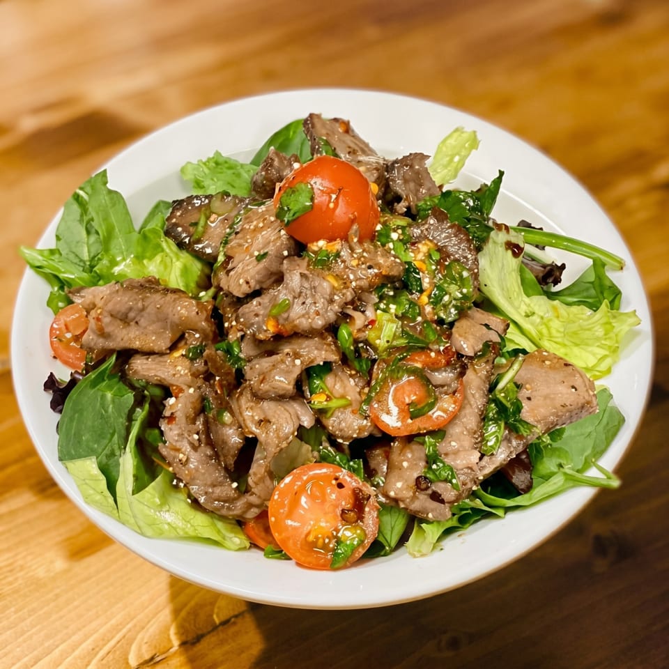 A white bowl filled with a beef salad, greens, and cherry tomatoes on a wooden table.