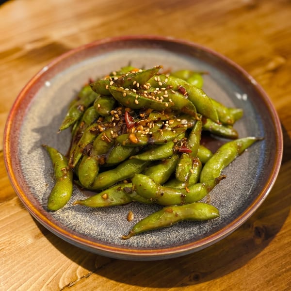 A plate of green edamame pods, seasoned with sesame seeds and a dark sauce, on a wooden table.