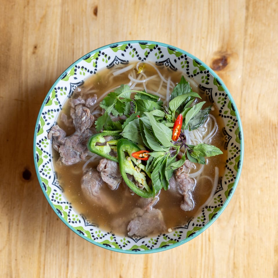 Overhead view of a colorful bowl of pho with beef, noodles, herbs, and chili peppers.