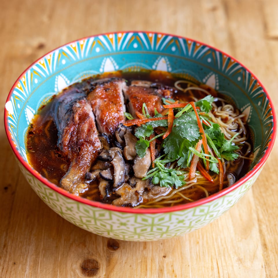 A colorful bowl of ramen with sliced duck, mushrooms, noodles, and fresh herbs on a wooden table.