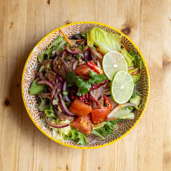 Spicy beef salad with red onions, tomatoes, chili, cilantro, and lime slices in a patterned bowl.