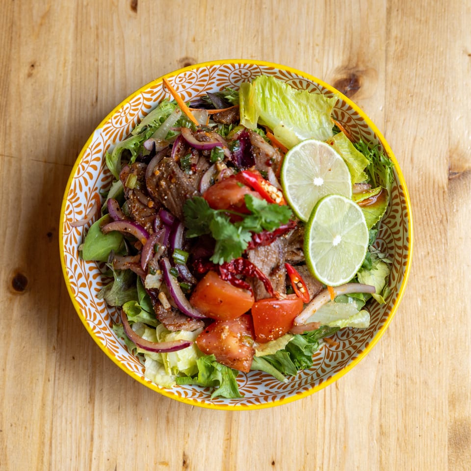 Spicy beef salad with red onions, tomatoes, chili, cilantro, and lime slices in a patterned bowl.