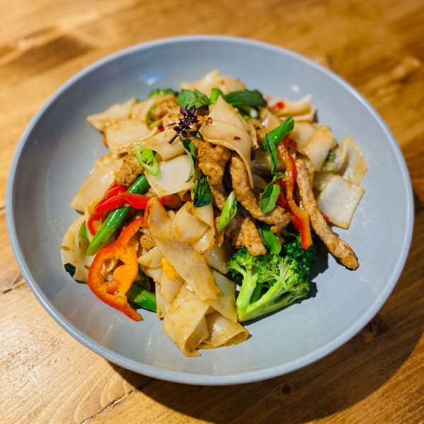 Close-up of a colorful Thai noodle dish with meat, broccoli, peppers, and basil in a blue bowl.
