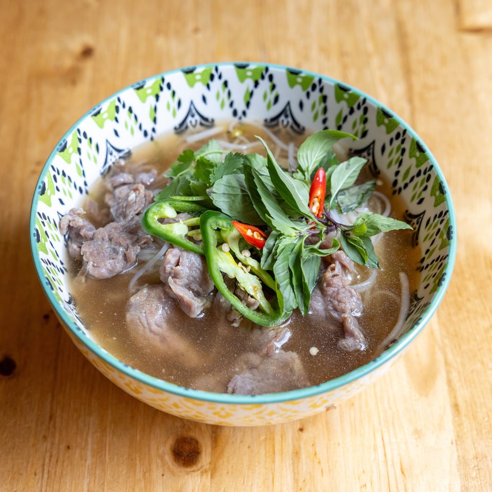 A colorful bowl of pho with beef, noodles, broth, basil, cilantro, and sliced chili peppers.