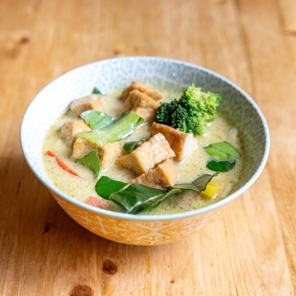 Green curry with tofu, broccoli, and kaffir lime leaves in a patterned bowl on a wooden table.