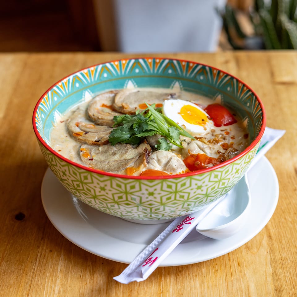 A colorful bowl of ramen with sliced pork, a halved egg, cilantro, and red peppers on a wooden table.